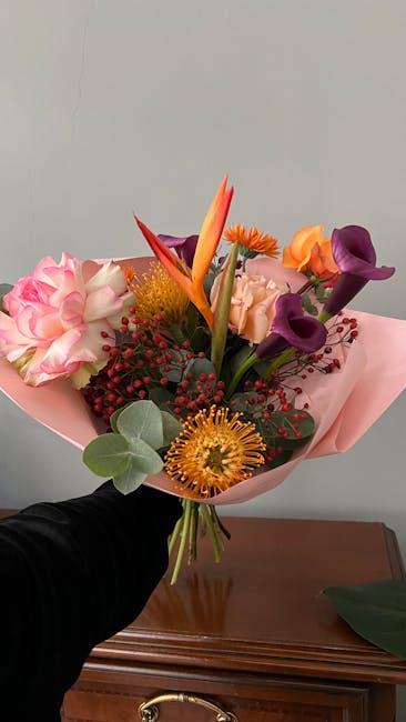 A vibrant bouquet of fresh flowers held by a hand against a neutral gray wall. The arrangement features a mix of blooms including pink roses, purple calla lilies, orange and purple tulips, a striking 