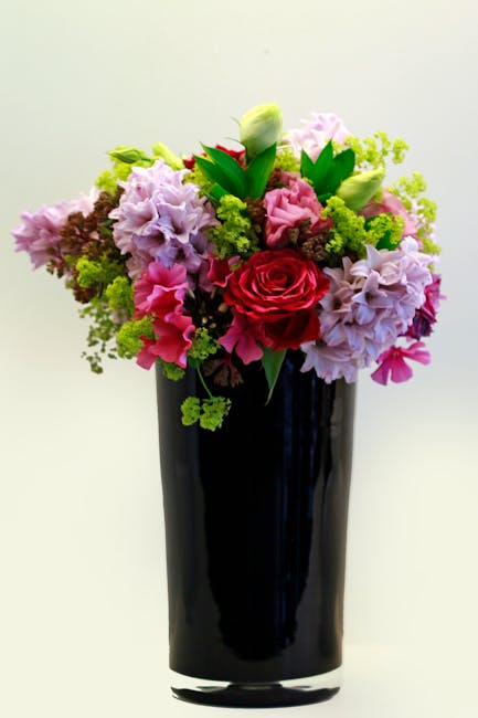A vibrant floral bouquet arranged in a tall, sleek black vase, featuring a mix of fresh flowers including a prominent deep red rose at the center, surrounded by soft pink hydrangeas, white tulips, and